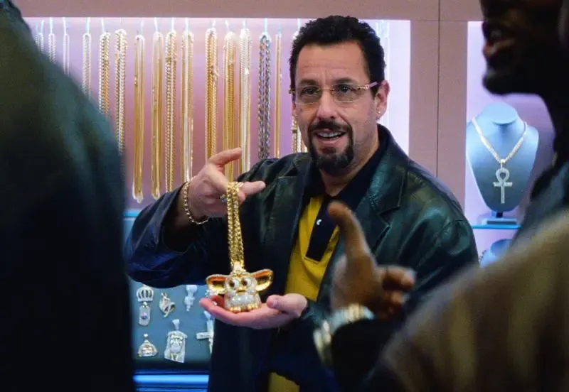 Adam Sandler as a jeweler in Uncut Gems, grinning as he holds up a gaudy gold Furby pendant on a chain in a display case full of gold jewelry.